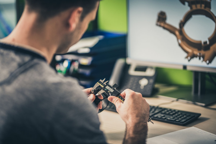 Over-the-shoulder view of a structural engineering student measuring a machine part while working on a 3D model of it.